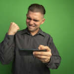 young handsome man grey shirt using gadget clenching fist happy excited rejoicing his success standing green wall