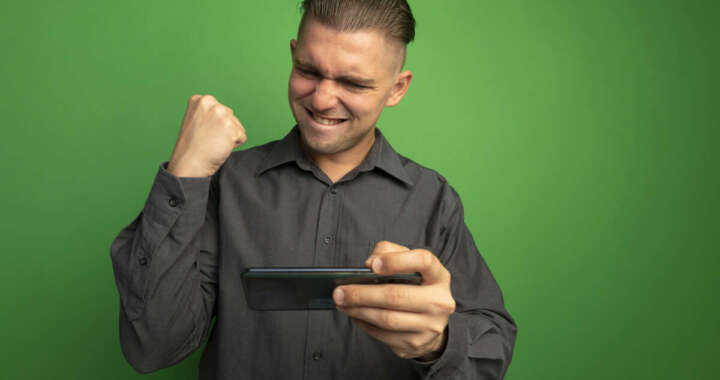 young handsome man grey shirt using gadget clenching fist happy excited rejoicing his success standing green wall